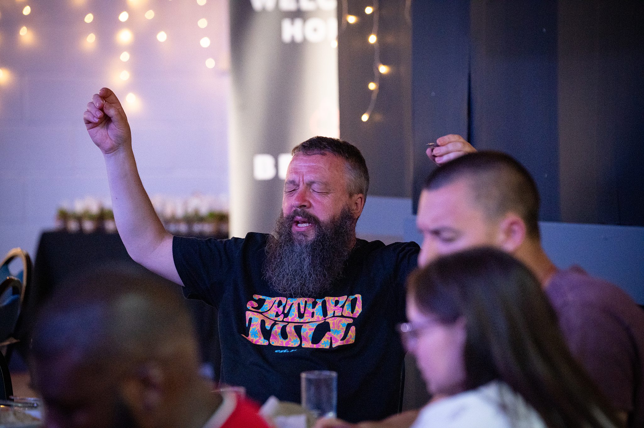 A man praying with raised hands during a church gathering in Bury St Edmunds, Suffolk, with people worshipping together in a warm community setting.