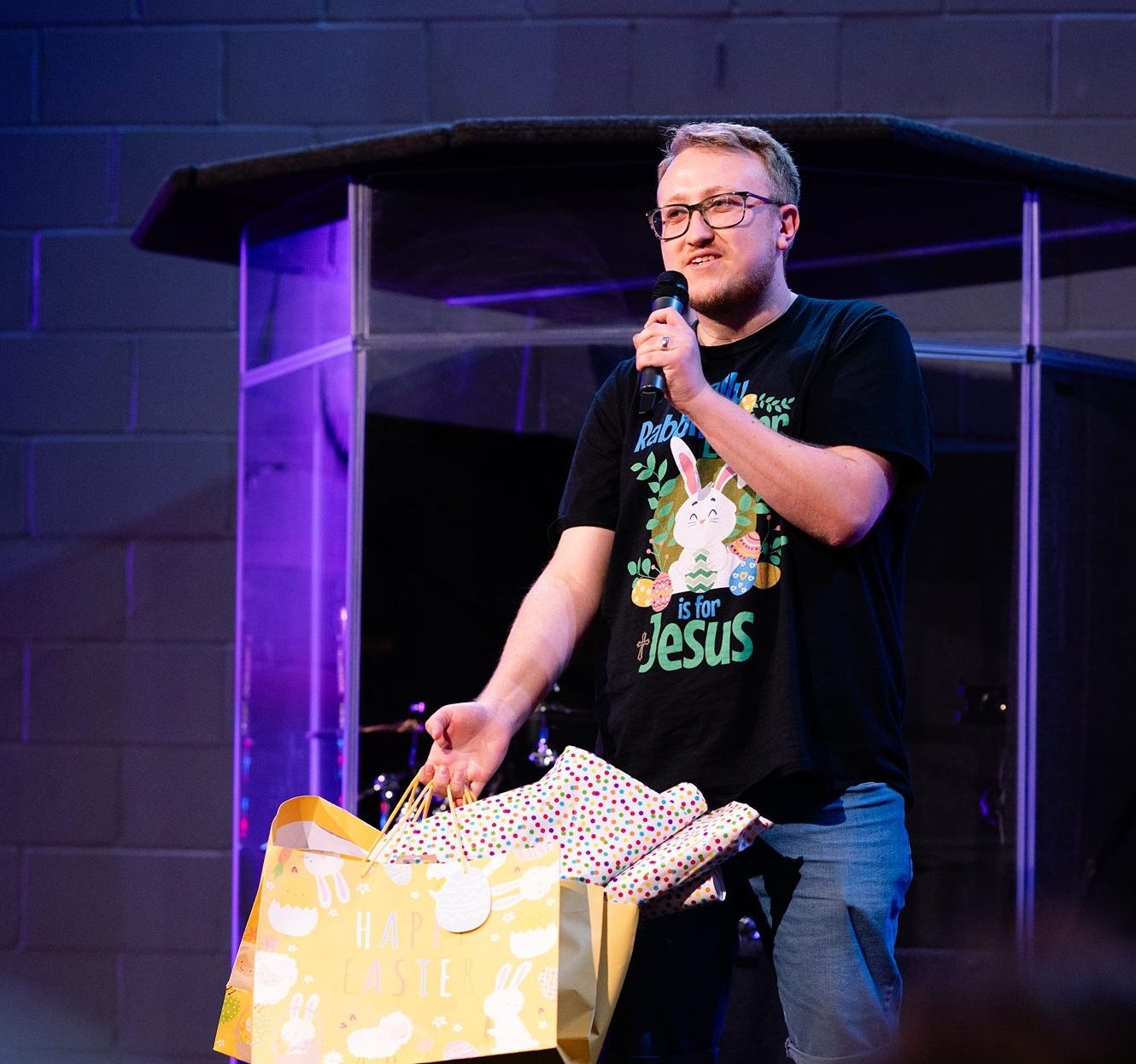 Man speaking on stage holding Easter gift bags during a church service in Bury St Edmunds, Suffolk