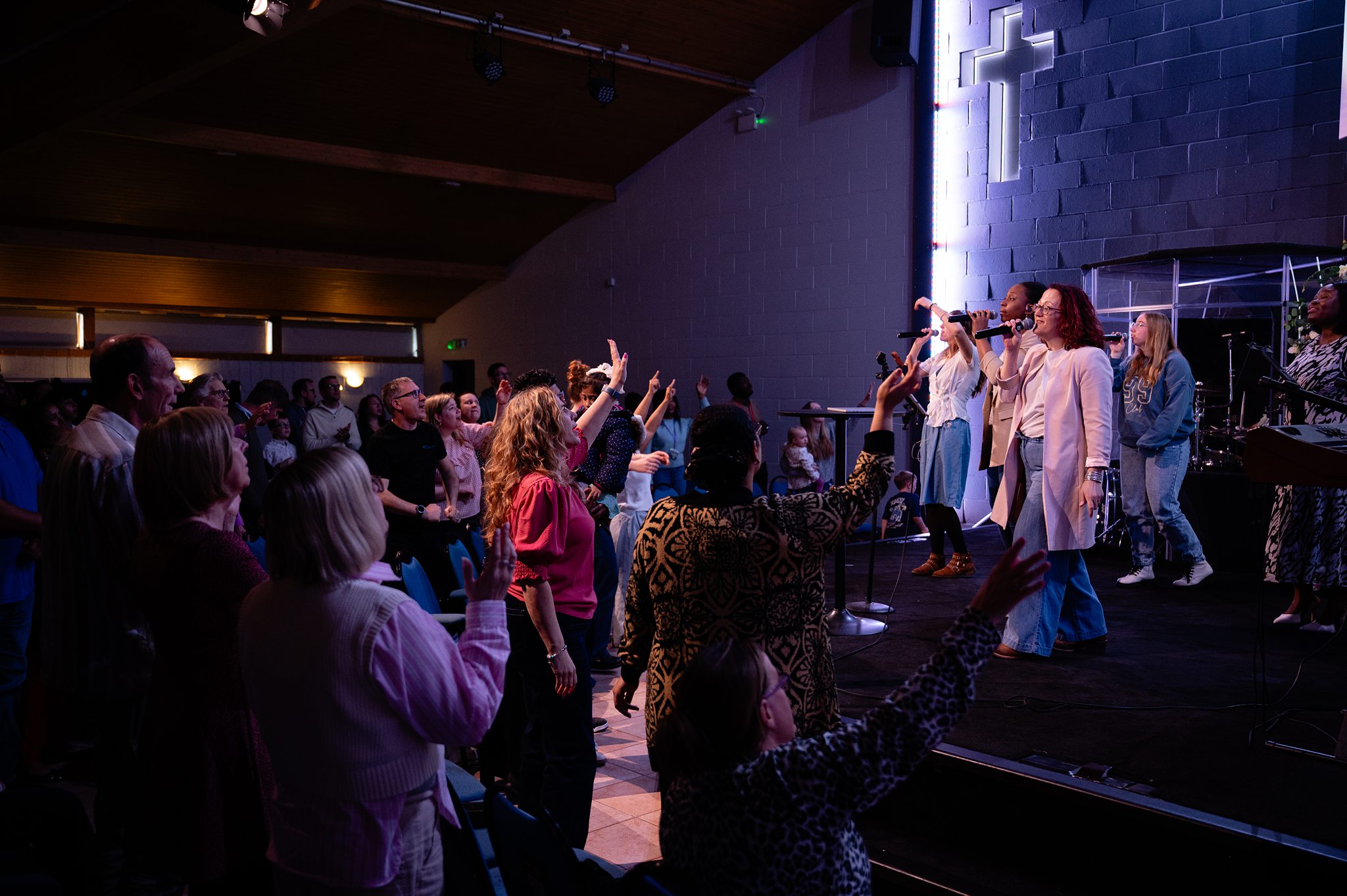 Congregation worshipping during a Sunday service at Beacon Church in Bury St Edmunds, Suffolk, with the worship team leading praise and worship as people raise their hands in prayer and celebration.